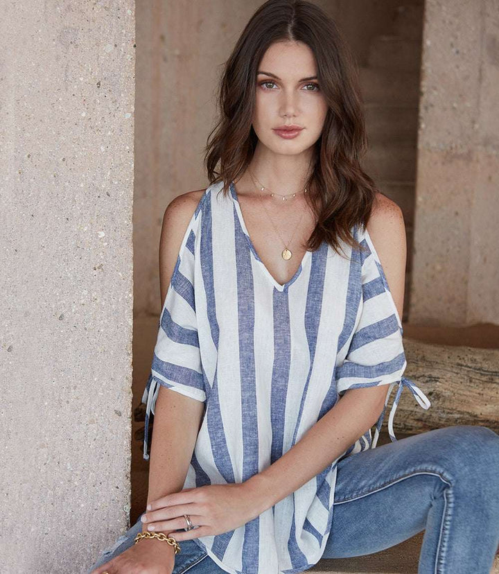 Woman wearing a blue and white striped cold shoulder tie sleeve top with a V-neck and shirttail hem.